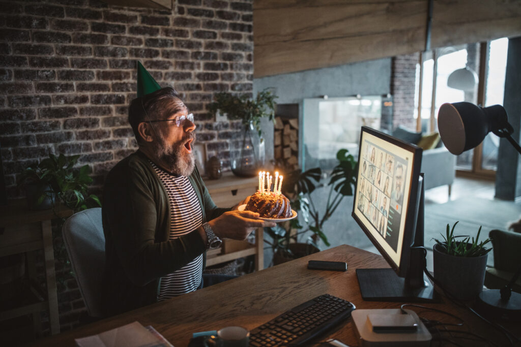 An Elderly man at home during pandemic isolation having a birthday celebration, he blowing candles and has a conference call with friends and family